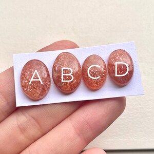 Peut inclure: Quatre cabochons ovales en pierre de soleil, &eacute;tiquet&eacute;s A, B, C et D, sur une carte blanche.