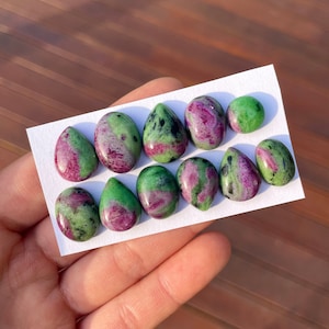 May include: A collection of ten teardrop-shaped green and pink gemstones. The stones are arranged on a white card.