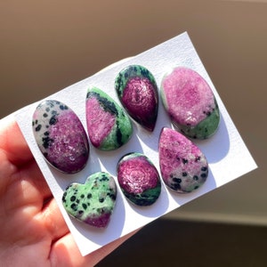 May include: A collection of seven green and pink gemstone cabochons, including a heart-shaped cabochon. The cabochons are set on a white background.