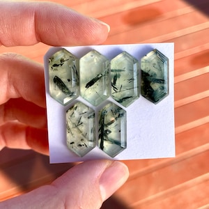 K&ouml;nnte beinhalten: Sechs sechseckige Edelsteine mit hellgr&uuml;ner Farbe und dunkelgr&uuml;nen Einschl&uuml;ssen. Die Edelsteine werden auf einer wei&szlig;en Karte pr&auml;sentiert, die von einer Hand gehalten wird. Der Hintergrund ist in einem verschwommenen Orange gehalten.