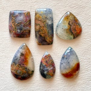 May include: Six polished gemstones in various shapes: rectangular, teardrop, and triangular. The stones display a mix of colors, including red, orange, gray, and brown, with unique patterns and textures. The gemstones are displayed on a light-colored surface.