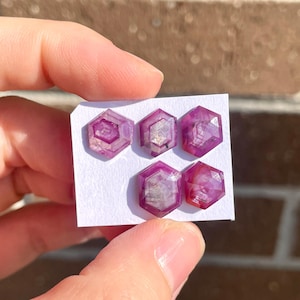May include: Five faceted pink sapphire gemstones on a white card. The gemstones are hexagonal in shape and have a faceted design.