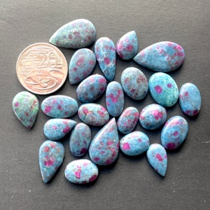 以下が含まれることがあります： 涙の形をした20個の青とピンクのルビーカボションの集まり。石は暗い灰色の表面に散らばっています。