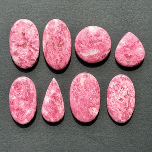 May include: Eight polished cabochons of pink rhodonite with white and black inclusions. The collection includes four oval shapes, two teardrop shapes, and two circular shapes, all displaying a smooth, glossy surface with natural stone patterns.