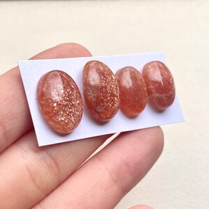 Peut inclure: Quatre cabochons ovales en pierre de soleil, chacun avec une couleur rouge-orange et une finition chatoyante et iris&eacute;e. Les pierres sont dispos&eacute;es en rang&eacute;e sur une carte blanche.
