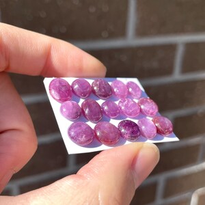 Pode incluir: Uma cole&ccedil;&atilde;o de pedras preciosas ovais polidas em tons de rosa e roxo. As pedras s&atilde;o dispostas em um cart&atilde;o branco, mostrando suas superf&iacute;cies lisas e varia&ccedil;&otilde;es naturais de cor. As pedras s&atilde;o seguradas por uma pessoa.