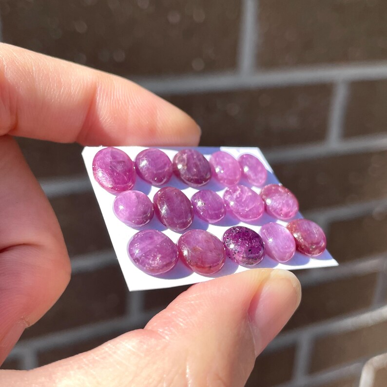 Pode incluir: Uma cole&ccedil;&atilde;o de pedras preciosas ovais polidas em tons de rosa e roxo. As pedras s&atilde;o dispostas em um cart&atilde;o branco, mostrando suas superf&iacute;cies lisas e varia&ccedil;&otilde;es naturais de cor. As pedras s&atilde;o seguradas por uma pessoa.