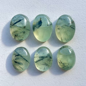 May include: Six oval prehnite gemstones. The stones are a translucent pale green with dark, needle-like inclusions. The polished stones are smooth and reflect light, showcasing their natural patterns and colours. These gemstones are ideal for jewellery making.