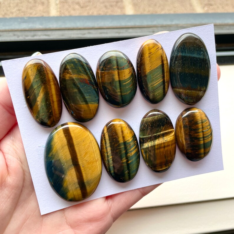 May include: Eight oval-shaped tiger's eye cabochons in various shades of brown, gold, and blue. The stones have a chatoyant effect, meaning they shimmer and change color in the light.