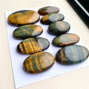 May include: Ten oval-shaped tiger's eye gemstones with a mix of golden brown and blue-green colors. The stones are polished and have a smooth, glossy finish.