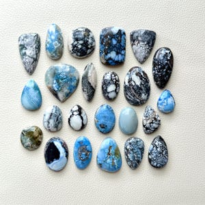 May include: A selection of polished gemstones in diverse shapes, such as oval, teardrop, and rectangular. The stones display a mix of blue, white, and black patterns, with some showing a marbled effect. The gemstones are arranged on a white surface.