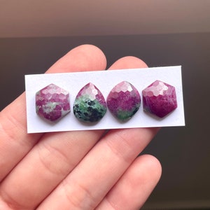 May include: Four faceted gemstones in shades of pink, green, and black. The stones are arranged in a row on a white card.