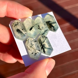 K&ouml;nnte beinhalten: Sechs hexagonale Prehnitkristalle, hellgr&uuml;n mit schwarzen Einschl&uuml;ssen, sind auf einer wei&szlig;en Oberfl&auml;che angeordnet. Die Kristalle werden in einer Hand gehalten. Die Prehnitkristalle sind durchscheinend.