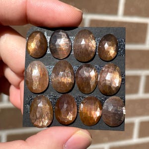 May include: A collection of 10 oval-shaped sunstone cabochons, each with a unique pattern of iridescent shimmer. The stones are arranged in two rows on a black background.