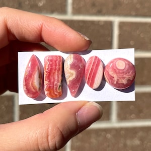 May include: Five pink and white gemstone cabochons, cut in various shapes, including oval, teardrop, and rectangular, displayed on a white card.