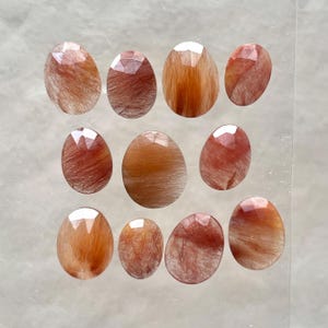 May include: Twelve oval-shaped, translucent, red and orange, gemstone cabochons with a hairy, fibrous texture.