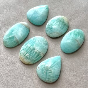 May include: Six polished Amazonite gemstones in various oval and teardrop shapes. The stones display a light blue-green color with white and tan veining patterns. The gemstones are arranged on a neutral-toned surface.