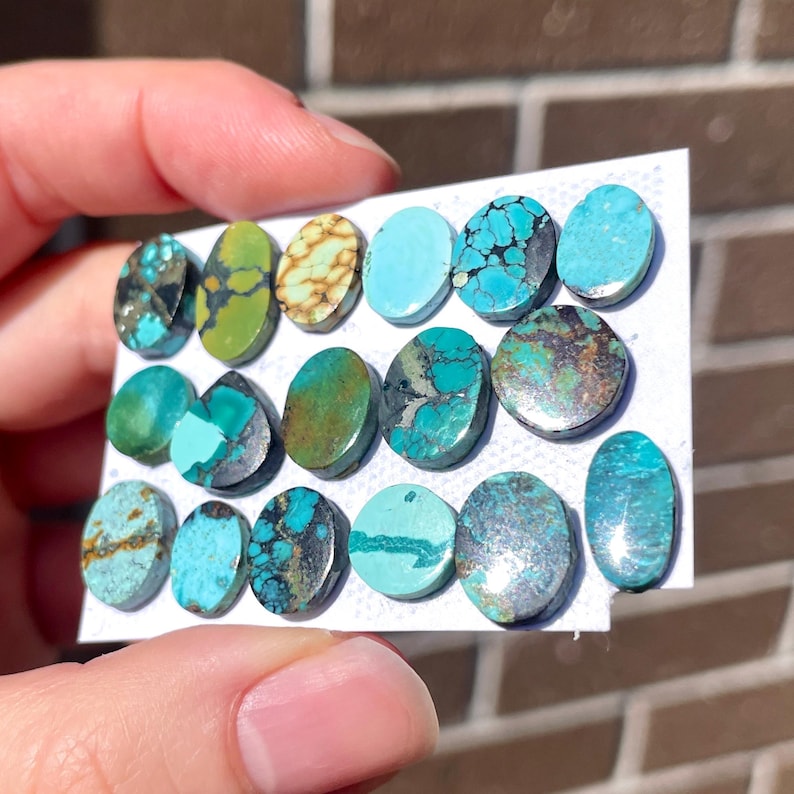May include: A collection of oval-shaped turquoise gemstones in various shades of blue, green, and brown. The stones are arranged on a white card, showcasing their unique patterns and textures. The gemstones are held by a hand.