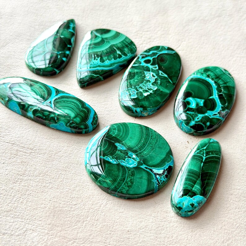 Pu&ograve; includere: Una collezione di sei pietre preziose di malachite lucidata in varie forme, tra cui goccia, ovale e circolare. Ogni pietra presenta tonalit&agrave; verde scuro con intricati motivi a bande vorticose e una finitura superficiale liscia.