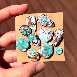 May include: A collection of turquoise gemstones in various shapes, including oval, teardrop, and heart. The stones feature a vibrant turquoise color with brown and white matrix patterns. Held against a brown background.