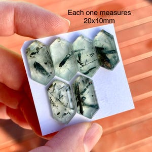 K&ouml;nnte beinhalten: Sechs durchscheinende, sechseckige Kristalle mit gr&uuml;nen und schwarzen Einschl&uuml;ssen. Jeder Kristall misst 20x10mm. Die Kristalle werden auf einer wei&szlig;en Oberfl&auml;che von einer Hand gehalten.
