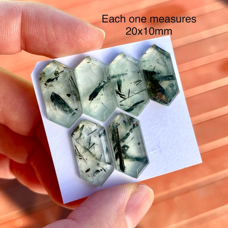 K&ouml;nnte beinhalten: Sechs durchscheinende, sechseckige Kristalle mit gr&uuml;nen und schwarzen Einschl&uuml;ssen. Jeder Kristall misst 20x10mm. Die Kristalle werden auf einer wei&szlig;en Oberfl&auml;che von einer Hand gehalten.