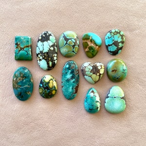 Op de afbeelding: Een verzameling van twaalf turquoise cabochons in verschillende vormen en maten, waaronder ovaal, rond, hartvormig en vierkant. De stenen zijn een mix van blauwe, groene en bruine kleuren.