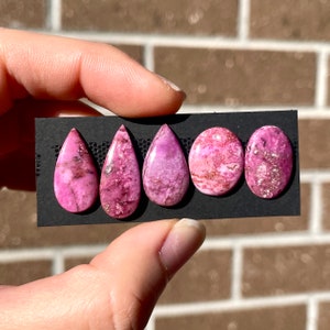 May include: Five pink and white cabochon gemstones in various teardrop and oval shapes, displayed on a black card.