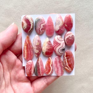 May include: A collection of 14 pink and white gemstone cabochons, each with a unique pattern of stripes and swirls. The cabochons are shaped like teardrops and are arranged in rows on a white surface.