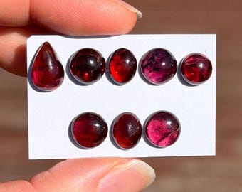 Pode incluir: Uma coleção de oito pedras preciosas polidas de cor vermelho profundo dispostas num cartão branco. O conjunto inclui uma pedra em forma de pera e sete cabochões redondos ou ovais, cada um exibindo níveis variados de translucidez.