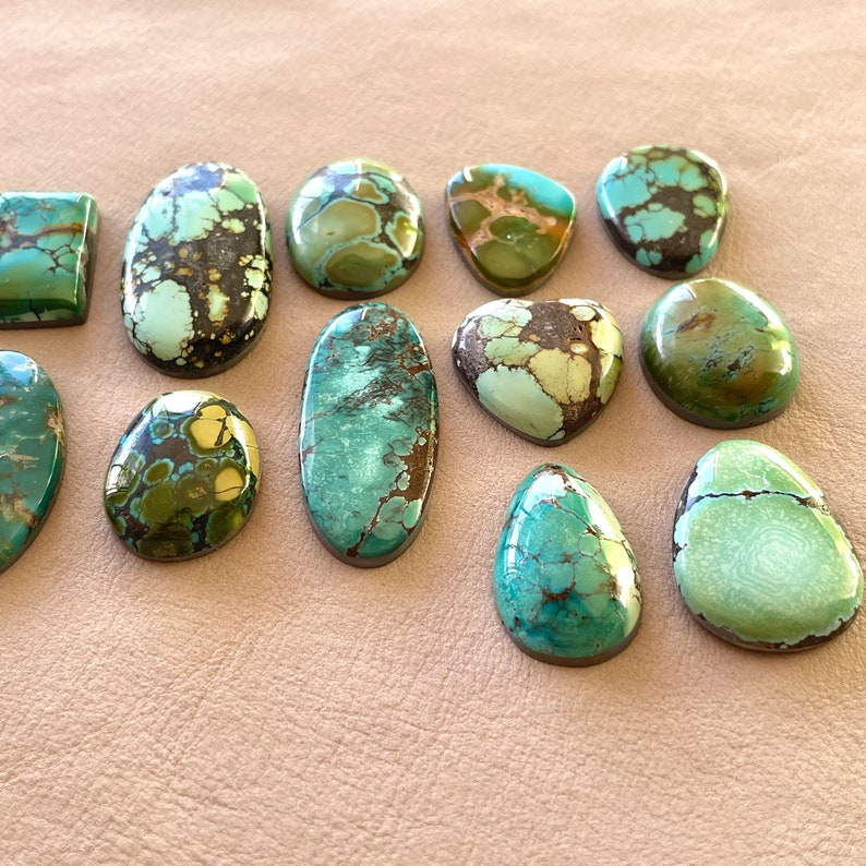 Op de afbeelding: Een verzameling van twaalf turquoise cabochons in verschillende vormen en maten. De stenen zijn een mix van groene, blauwe en bruine kleuren met een marmerpatroon.