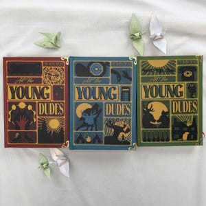 May include: Three hardcover books with the title "All The Young Dudes" in gold lettering. The books are in red, blue, and green, each with unique illustrations and gold corner accents. Origami cranes are placed around the books.