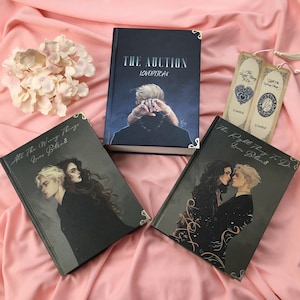 May include: A collection of books and bookmarks with dark covers and gold corner accents. The books feature illustrations of couples and the titles "The Auction" and "All The Wrong Things Love Blends". Bookmarks with text are also present.