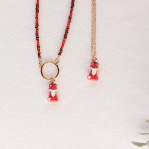 Könnte beinhalten: Eine Halskette mit einer Reihe kleiner roter und brauner Perlen, einem goldfarbenen Ring und einem kleinen rot-weißen Fuchs-Anhänger. Daneben hängt eine zweite, dünnere goldfarbene Kette, die ebenfalls in einem passenden Fuchs-Anhänger endet.