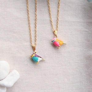 Pode incluir: Duas correntes douradas com pingentes de pássaros. Um pingente é azul, turquesa, branco e roxo. O outro é rosa, laranja e azul. Os pingentes de pássaros têm pés dourados e estão em uma superfície branca.