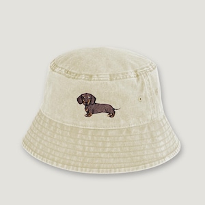 Peut inclure: Un chapeau bob beige avec un motif brodé d'un chien teckel marron. Le chapeau a un design décontracté et un aspect légèrement délavé. Le chien est détaillé avec des poils bruns et noirs.