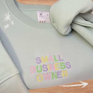 Op de afbeelding: Een lichtgroene sweatshirt met de geborduurde tekst "SMALL BUSINESS OWNER" in kleurrijke letters. De sweatshirt heeft een ronde hals en een label met "B&C Collection". Een witte pijl wijst naar rechts.