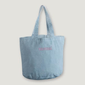 Könnte beinhalten: Eine hellblaue Tragetasche aus Denim mit zwei Schulterriemen und einem abgerundeten Boden. Das Wort mausi ist in kleinen rosa Buchstaben mittig auf der Vorderseite der Tasche eingestickt. Der Stoff hat eine sichtbare Webstruktur.