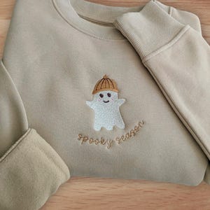 Könnte beinhalten: Ein beigefarbenes Sweatshirt mit Rundhalsausschnitt und einer Stickerei eines kleinen weißen Geistes, der eine braune Mütze trägt. Unter dem Geist ist der Text "spooky season" in brauner Schreibschrift mittig auf der Brust eingestickt.