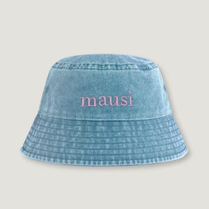 Könnte beinhalten: Ein blauer Fischerhut aus Denim mit verwaschener, abgenutzter Textur. Auf der Vorderseite des Hutes ist das Wort "mausi" in hellrosa, kleingeschriebenen Serifenschriftzeichen mittig auf der Krone über der Krempe eingestickt.