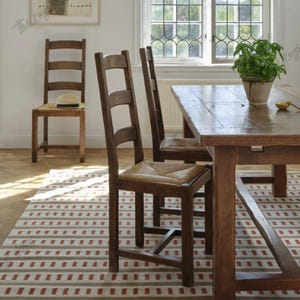 May include: A dining room scene featuring a wooden table and chairs. The chairs have woven seats. A rug with a red and white striped pattern is on the floor. A framed artwork hangs on the wall, and a plant sits on the table.