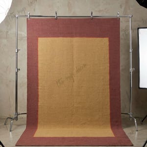 May include: A rectangular area rug featuring a large mustard yellow center panel framed by a wide border in a deep terracotta red. The rug is suspended from a metal stand with silver clips, displaying a flat-woven texture and a clean, simple design.
