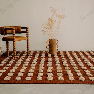 May include: A rectangular, russet-coloured rug with a repeating pattern of off-white tufts. A wooden chair with a rounded back and legs is to the left. A brown vase with dried flowers sits on the rug.