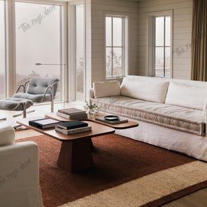 Puede incluir: Un salón con una alfombra marrón y beige, una mesa de centro de madera con libros y un sofá blanco. La habitación tiene grandes ventanales y una paleta de colores neutros, creando un ambiente acogedor y agradable.