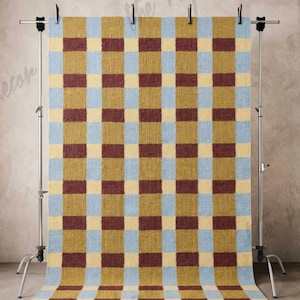 May include: A rectangular rug with a checkered pattern in shades of blue, gold, and burgundy. The rug is displayed on a metal stand with clips, showcasing its design and texture. The rug's pattern is a grid of alternating colors.