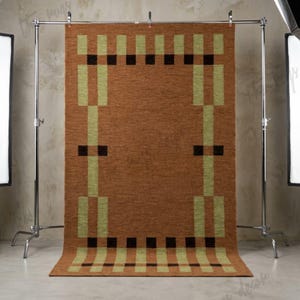 Puede incluir: Una alfombra rectangular marrón con un diseño geométrico. La alfombra presenta un patrón repetitivo de rectángulos y cuadrados verdes claros y negros. La alfombra se exhibe en un estante de metal en un estudio.