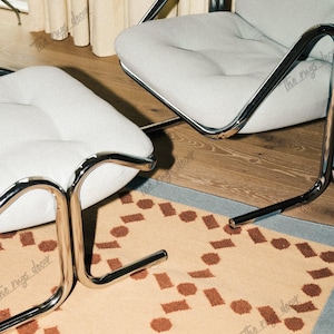 Pode incluir: Uma cena de sala de estar moderna com uma cadeira e um pufe estofados em cinza claro com estruturas cromadas. Um tapete bege com formas geométricas marrons está sob os móveis. A sala tem pisos de madeira e uma paleta de cores neutras.