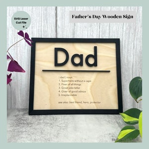 May include: Wooden sign with the word "Dad" in black lettering on a light wood background. The sign also includes a definition of the word "dad" with five bullet points and the phrase "see also: best friend, hero, protector".