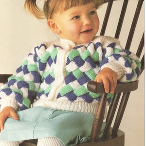 May include: A white knitted cardigan with a green, blue, and white geometric pattern. The cardigan has a button closure and is worn by a young child.
