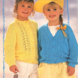 Könnte beinhalten: Zwei Kinder tragen gestrickte Cardigans. Das Mädchen links trägt einen gelben Cardigan mit Knopfleiste. Das Mädchen rechts trägt einen blauen Cardigan mit Knopfleiste. Beide Cardigans haben ein Spitzenmuster.